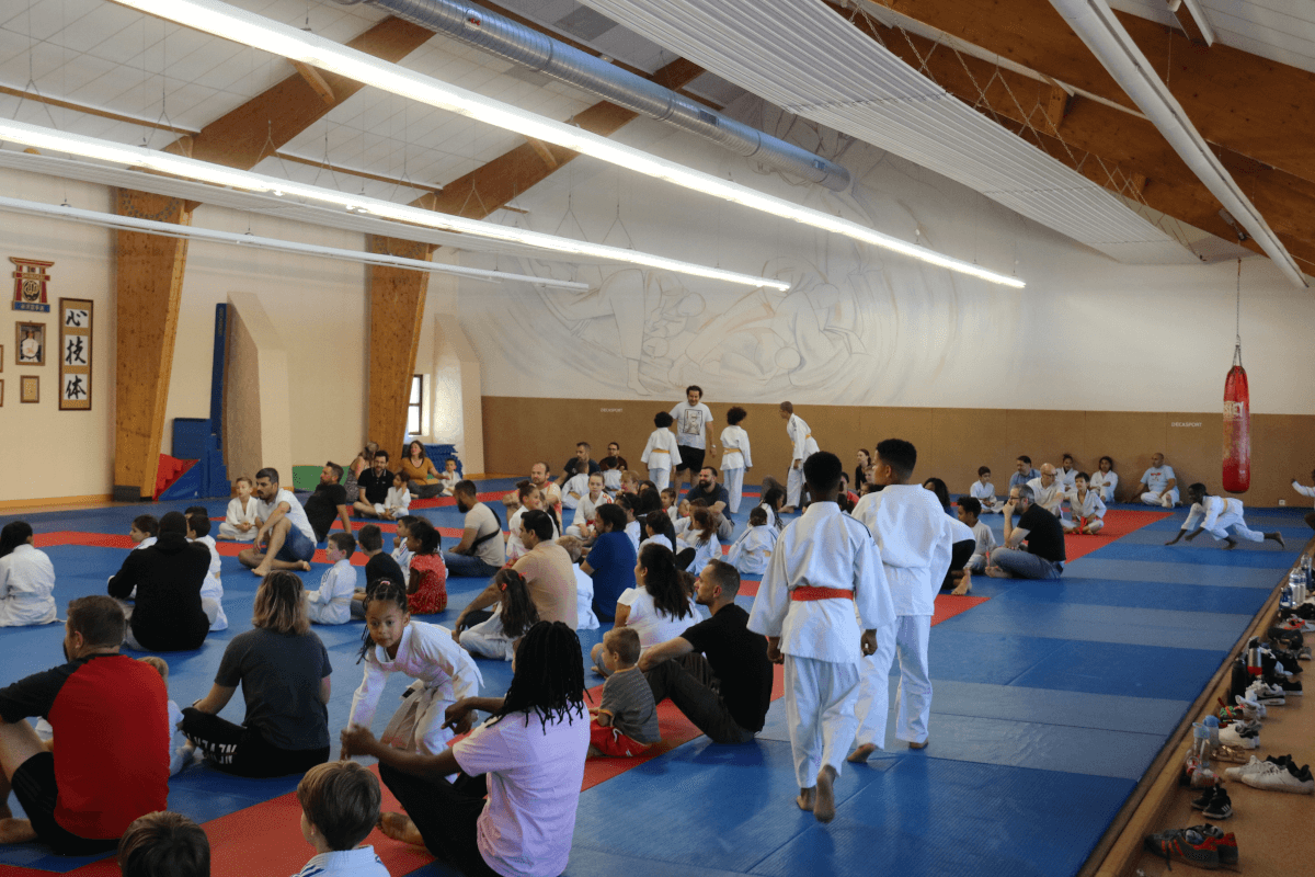 Un jeune garçon recevant sa nouvelle ceinture blanche de la part d'un instructeur, pendant que d'autres judokas et spectateurs regardent.