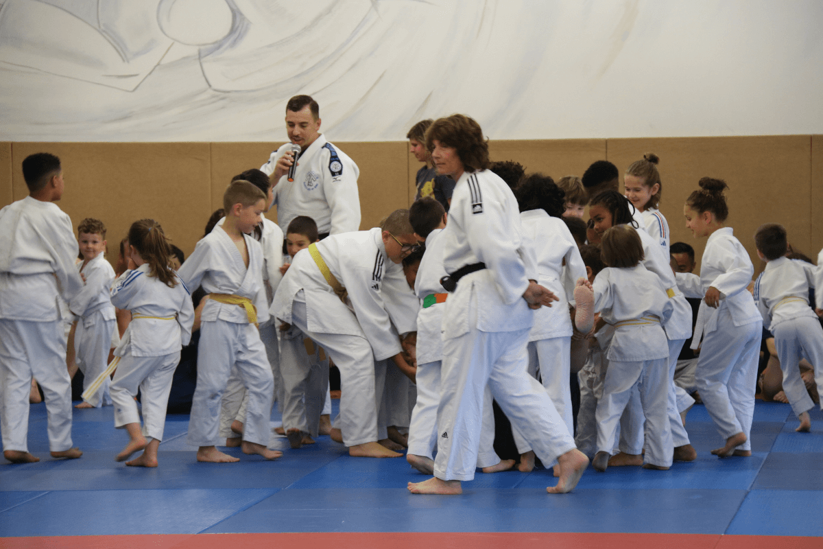 Deux judokas exécutant une technique de projection, sous le regard attentif de jeunes enfants assis au fond.