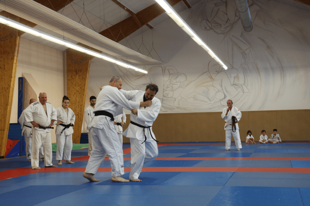 Démonstration de judo où un pratiquant ceinture noire exécute une projection sur un autre judoka, sous les regards d'enfants assis.