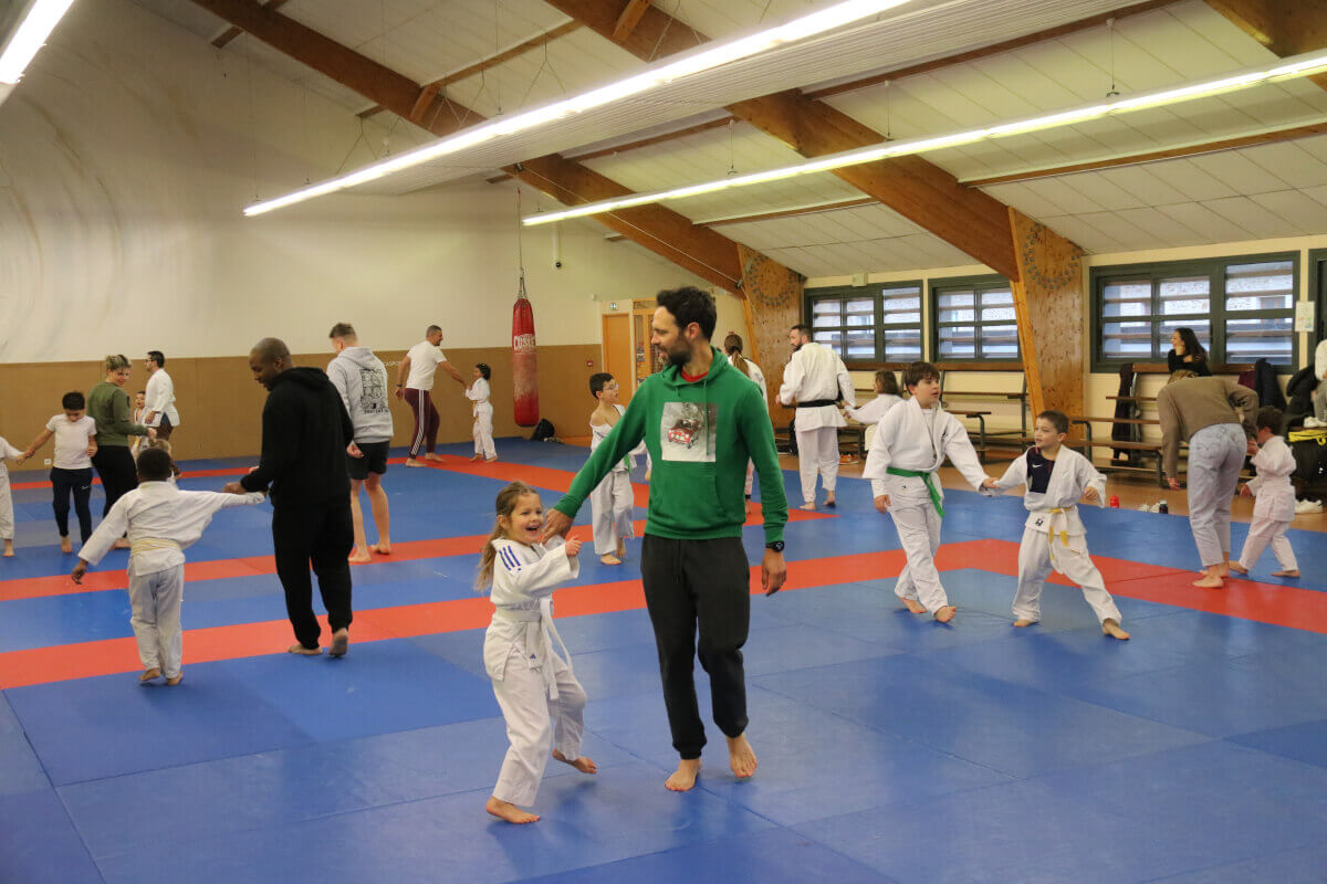 Un jeune garçon recevant sa nouvelle ceinture blanche de la part d'un instructeur, pendant que d'autres judokas et spectateurs regardent.