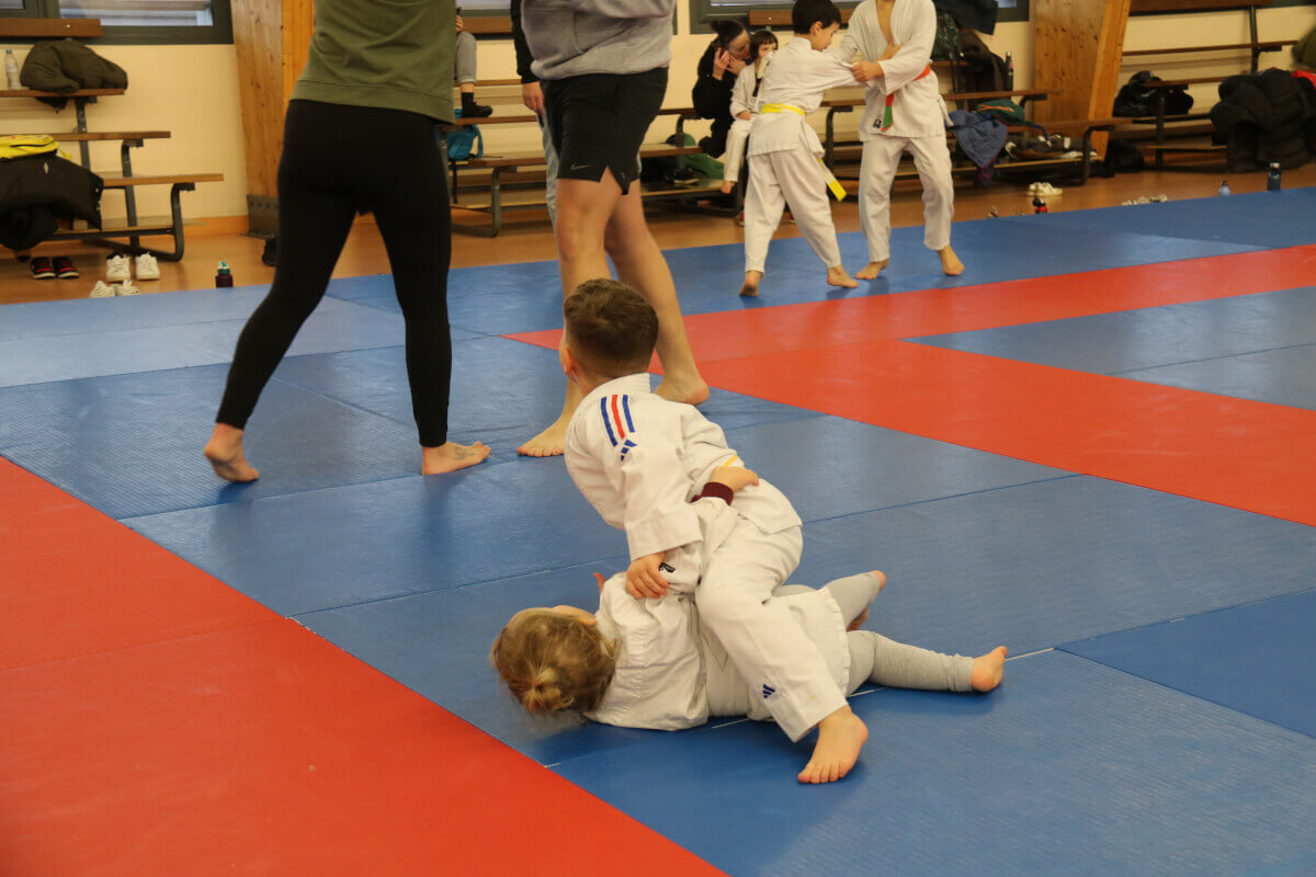 Démonstration de judo où un pratiquant ceinture noire exécute une projection sur un autre judoka, sous les regards d'enfants assis.