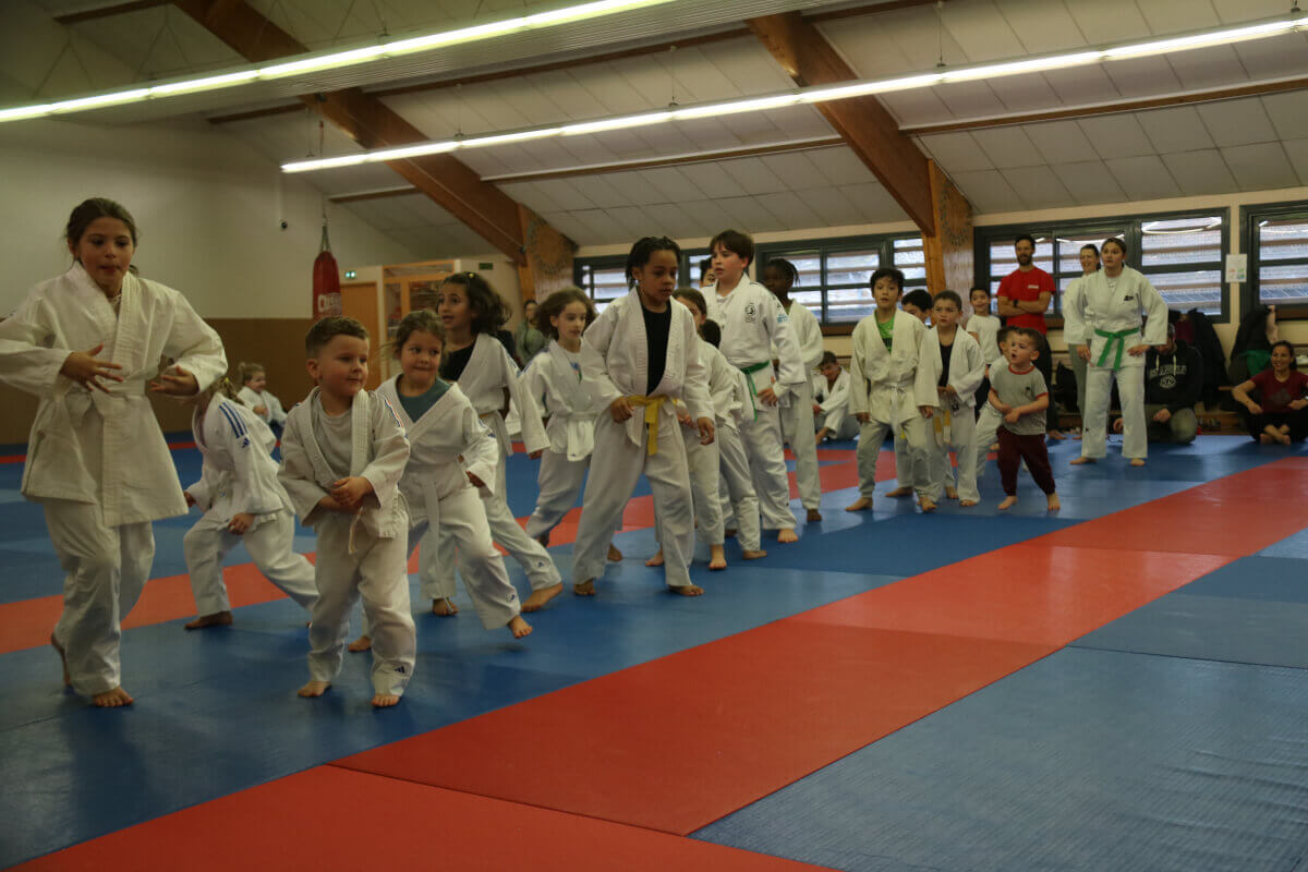 Des enfants alignés en tenue de judo, attendant leur tour pour une activité, avec des ceintures colorées exposées devant eux.