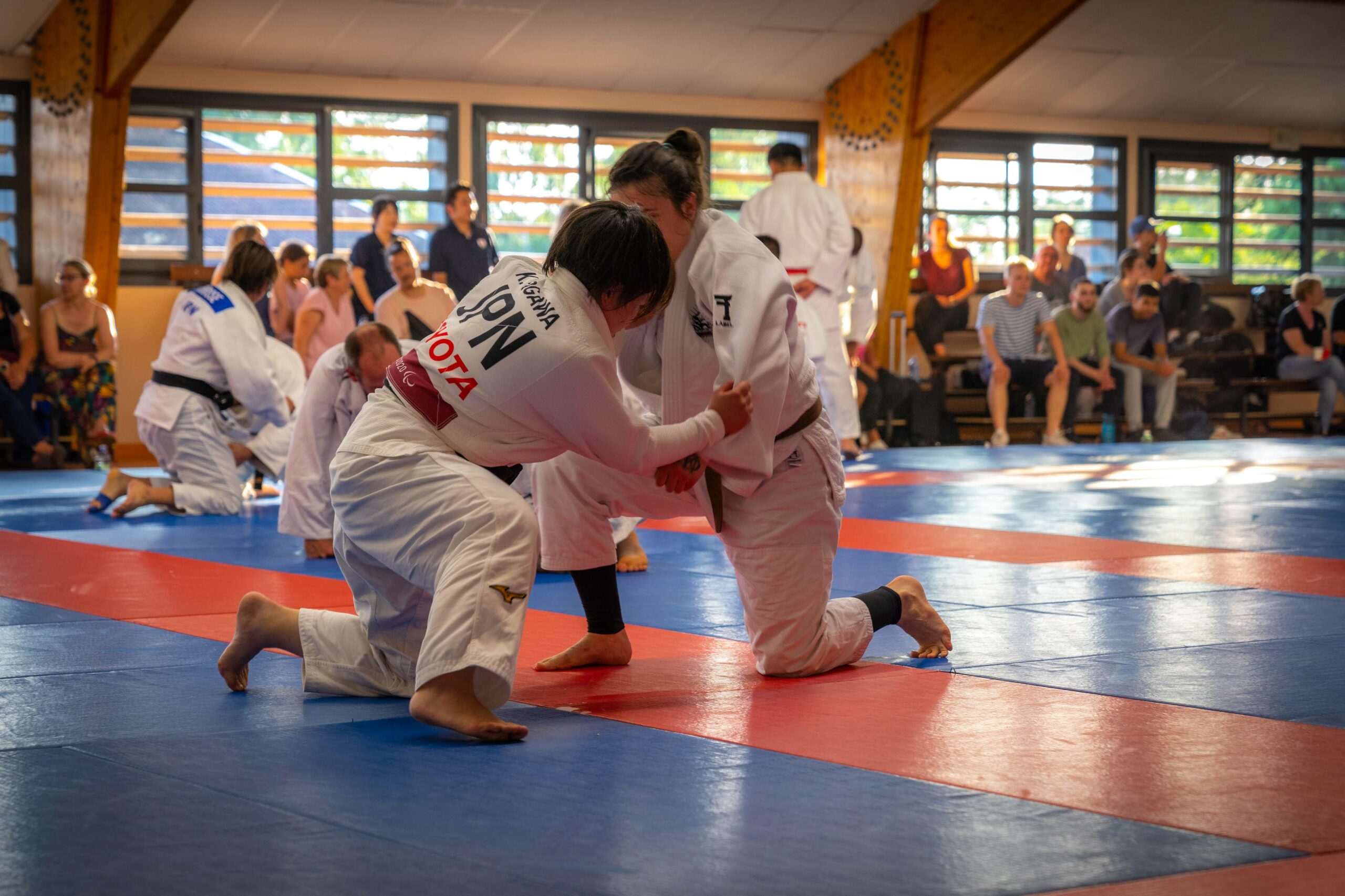 Deux judokas en plein exercice de lutte au sol, spectateurs autour.