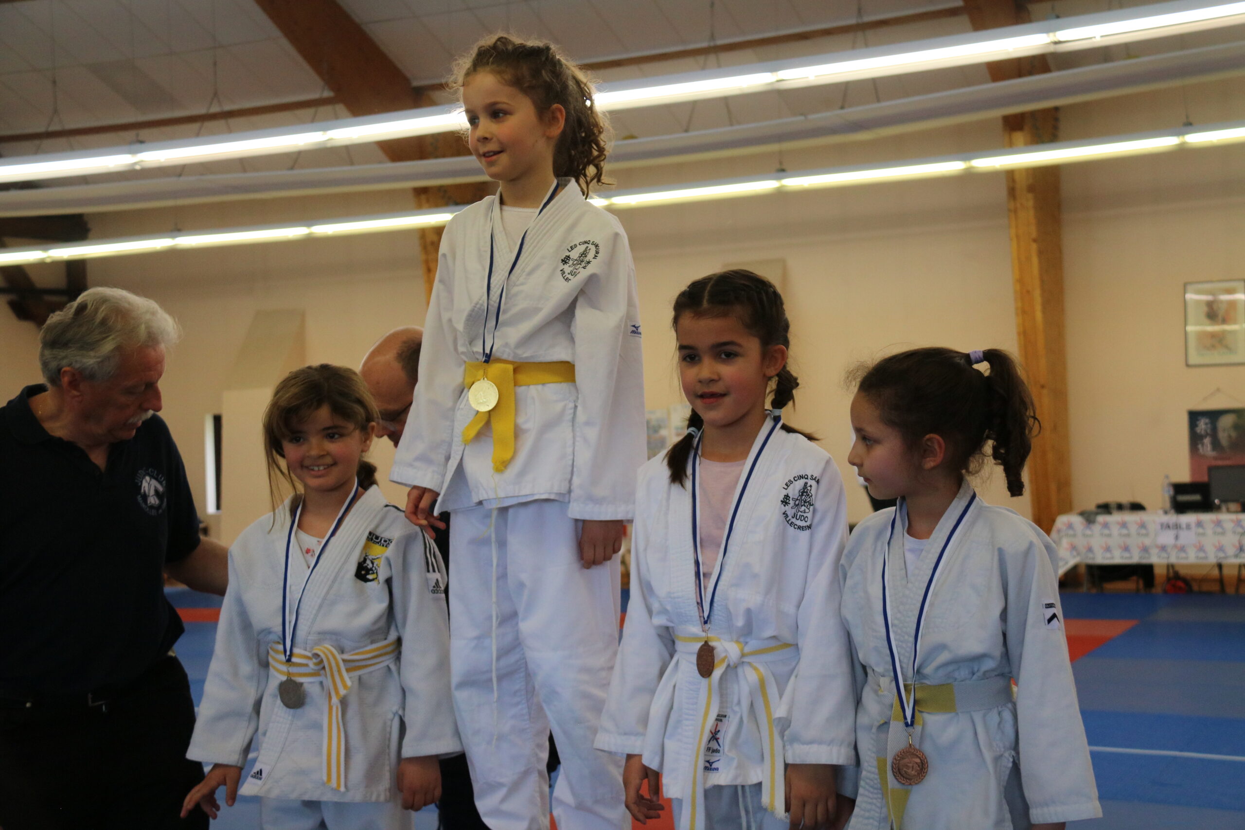 Podium avec quatre jeunes filles judokas récompensées lors d'un tournoi, avec l'entraîneur les félicitant.