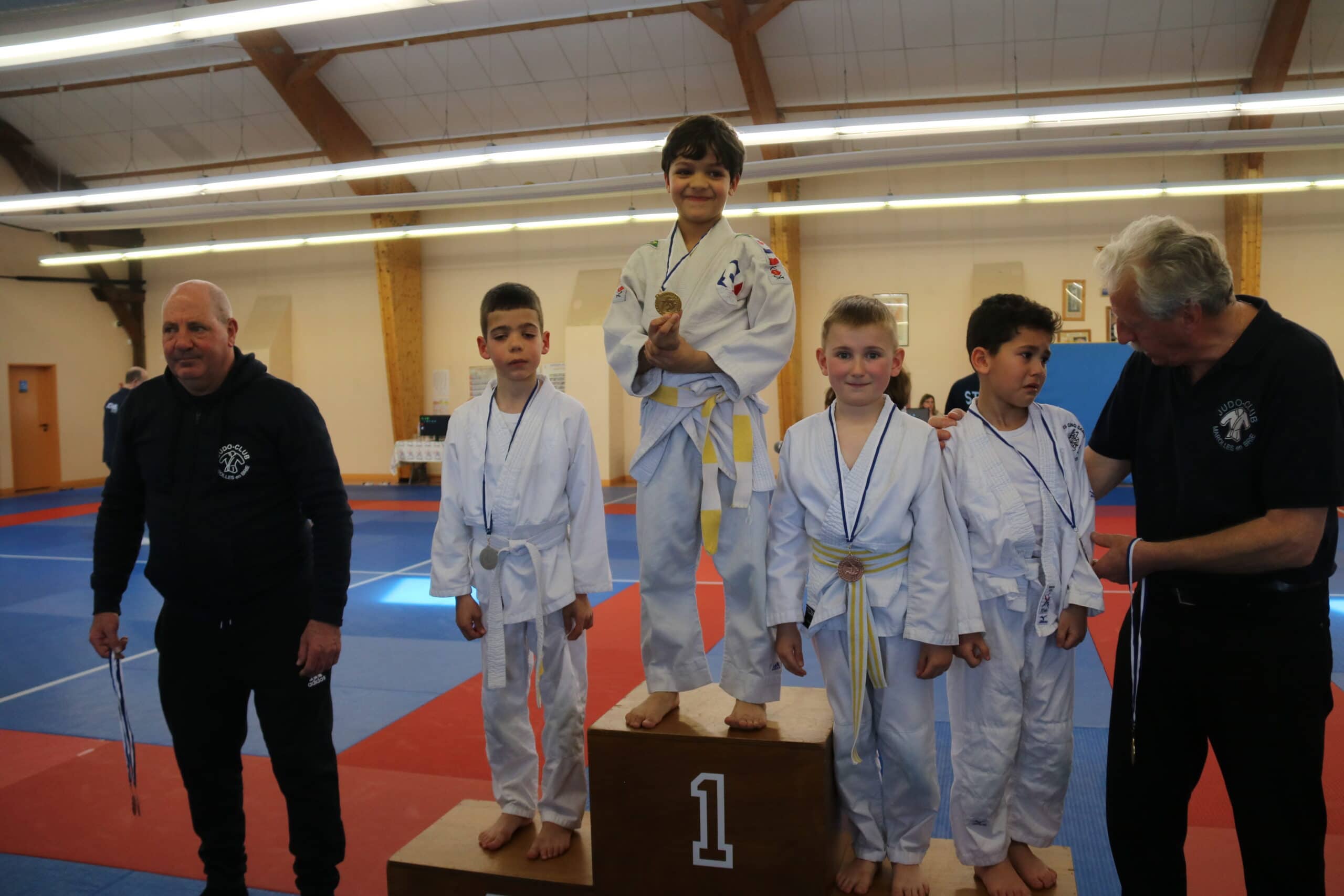 Podium de jeunes judokas avec leurs médailles, l'entraîneur les félicite après la cérémonie.