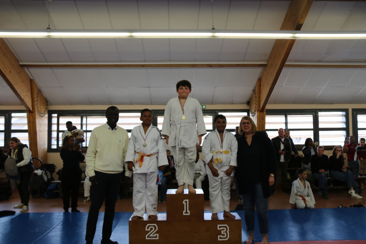 Podium avec quatre jeunes filles judokas récompensées lors d'un tournoi, avec l'entraîneur les félicitant.