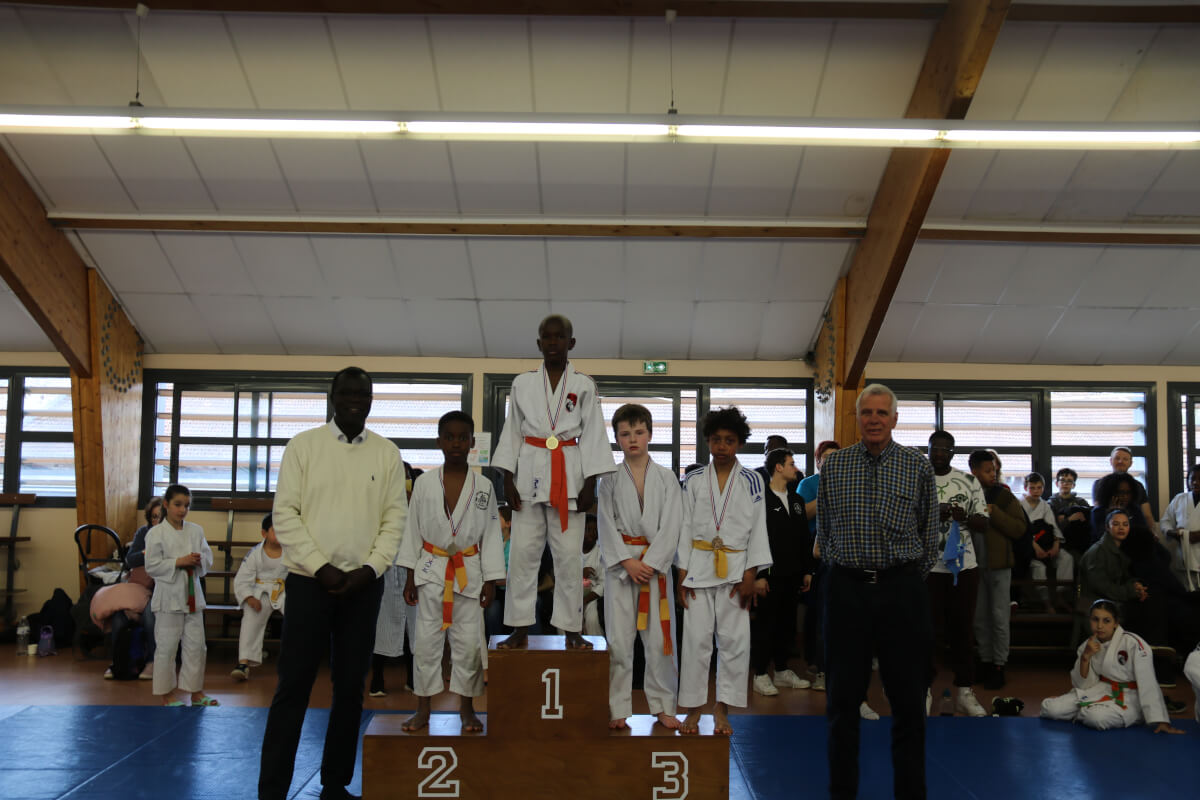 Podium de jeunes judokas avec leurs médailles, l'entraîneur les félicite après la cérémonie.